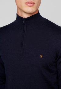 Mörkblå zip-neck-tröja med ribbat mönster. Har en liten orange logotyp på vänster bröst. Tätt passande design, mjukt tyg.