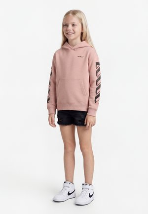 Smiling child with long blonde hair wearing a pink hooded sweatshirt with black stripes on sleeves, black ripped shorts, and white Nike sneakers.