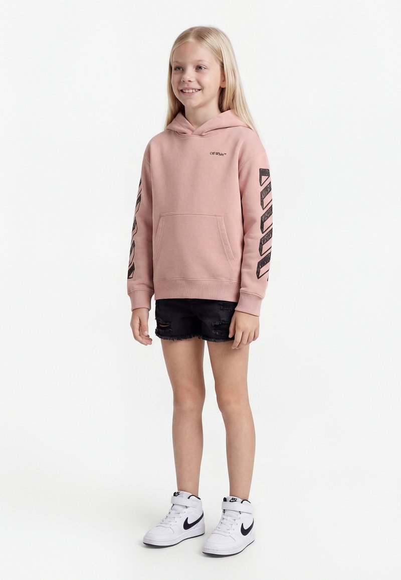 Smiling child with long blonde hair wearing a pink hooded sweatshirt with black stripes on sleeves, black ripped shorts, and white Nike sneakers.