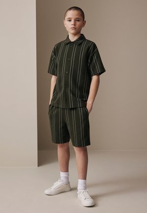 Boy standing with hands in pockets wearing dark green short-sleeve button shirt and matching shorts with white vertical stripes, white socks, and sneakers.