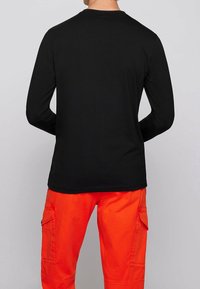 Schwarzes Langarmshirt mit Rundhalsausschnitt, getragen mit leuchtend orangefarbener Cargo-Hose mit Seitentaschen. Glatter Stoff, lockere Passform.
