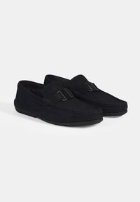 Schwarze Wildleder-Slipper mit flacher Sohle, dekorativem Riemen und Schnallendetail, gesteppten Akzenten und einem weichen Innenfutter.