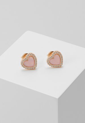 Boucles d’oreilles clous en forme de cœur avec centres roses entourés de petites pierres transparentes sur une surface d’exposition blanche.
