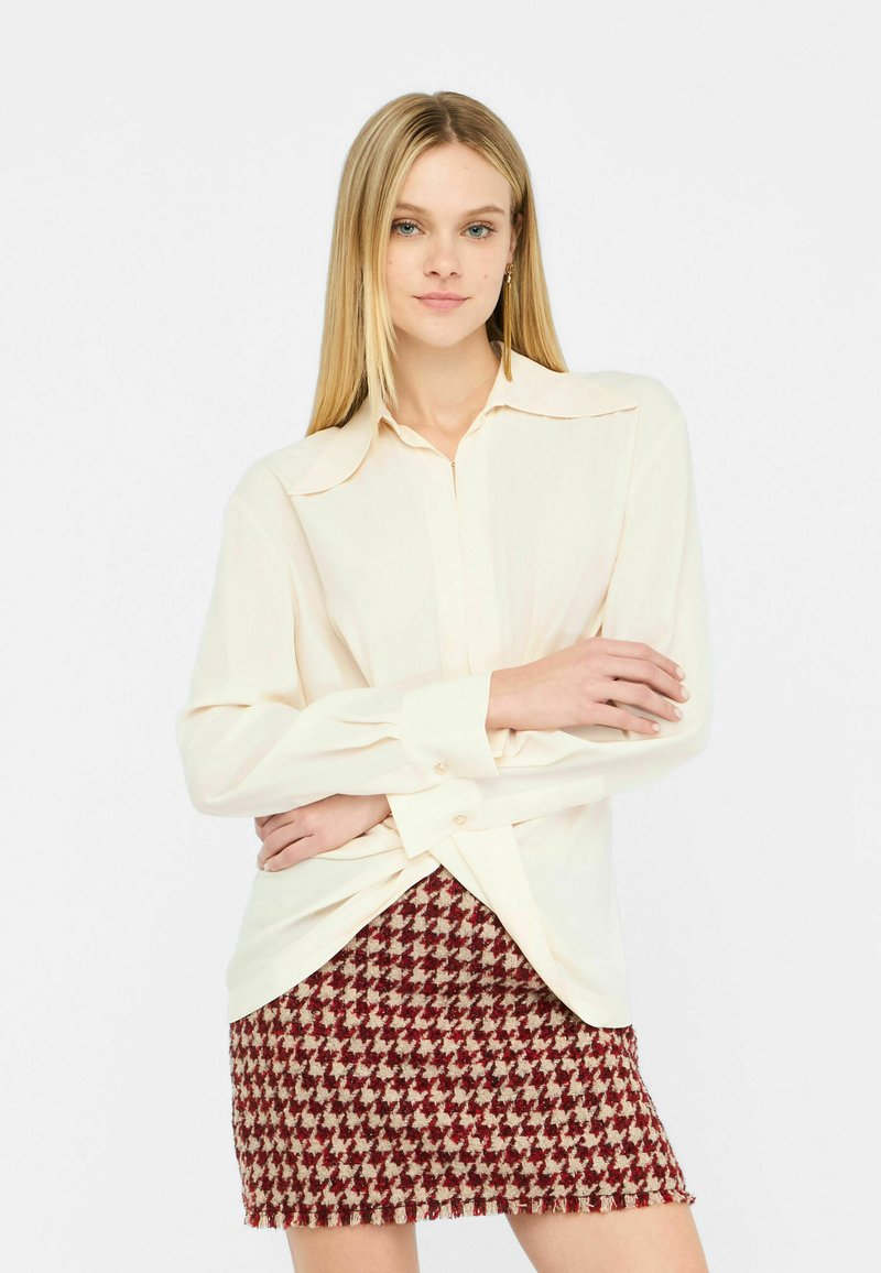 Camicia crema con colletto a pieghe e maniche lunghe, abbinata a una mini gonna in tessuto a scacchi rosso e beige con orlo sfrangiato.