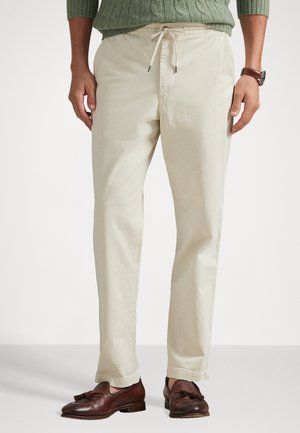 Smėlio spalvos medvilninės kelnės su elastingu juosmens diržu, raišteliu ir siaurėjančiomis kojomis; vilkimos su rudais odiniais loaferiais.