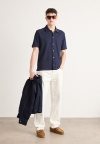 Camicia a maniche corte blu navy con bottoni, pantaloni bianchi, scarpe marroni, tiene in mano una giacca blu navy, occhiali da sole. Tessuto leggero, vestibilità moderna, stile casual.