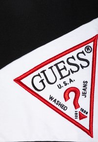 Rød og hvit brodert trekantlogo med "GUESS" og "WASHED JEANS" tekst på en svart stoffbakgrunn.