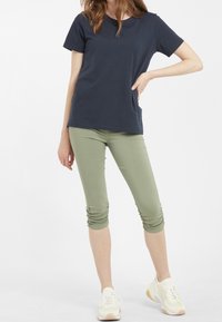 Námořnické tričko spárované s olivově zelenými 3/4 legínami. Legíny mají na bocích nařasené detaily, nošeny s bílými a žlutými sportovními botami.