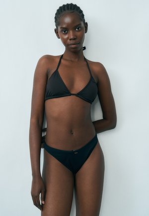 Junge Frau mit geflochtenem Haar, die einen schwarzen Bikini trägt, steht vor einer schlichten hellgrauen Wand und blickt direkt in die Kamera.