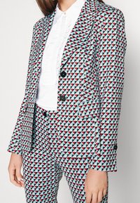 Costume sur mesure avec un motif géométrique de triangles bleus, rouges et noirs. Il comprend un revers cranté, deux boutons et un pantalon ajusté.