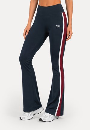 Femme portant un pantalon de sport bleu marine évasé avec des bandes latérales blanches et rouges, logo FILA sur la hanche, et des baskets blanches, vue de la taille vers le bas.