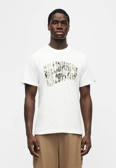 Νέος άνδρας φορώντας λευκό κοντομάνικο T-shirt με το κείμενο "Billionaire Boys Club" και καφέ παντελόνι, στέκεται μπροστά σε ένα απλό φωτεινό φόντο.