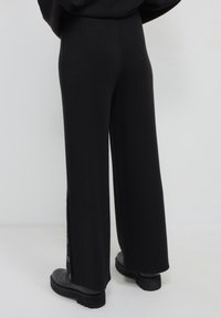 Person, der schwarze weite Hosen mit seitlichen Knöpfen am Saum trägt, kombiniert mit schwarzen Schuhen mit dicker Sohle, steht vor einem schlichten Hintergrund.