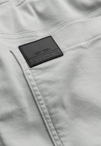 Tissu gris clair avec coutures visibles et une étiquette rectangulaire noire cousue, montrant un texte embossé et un marquage.
