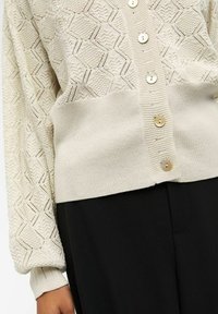Cárdigan de punto beige claro con un patrón geométrico, cuello redondo, mangas largas y botones de perla. Tejido texturizado y puños acanalados.