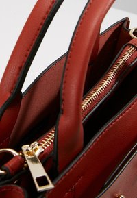 Sac à main en simili cuir rouge avec quincaillerie dorée, anses doubles et compartiments zippés. Présente une texture lisse et une garniture noire contrastante.