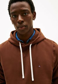 Felpa marrone con coulisse bianche, con una camicia color blu sotto. Dettaglio logo evidente sul petto. Texture morbida.