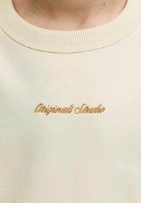 Sweatshirt bege claro com um colarinho redondo em canelado, apresentando texto bordado em dourado a dizer "Originals Studio" na frente.