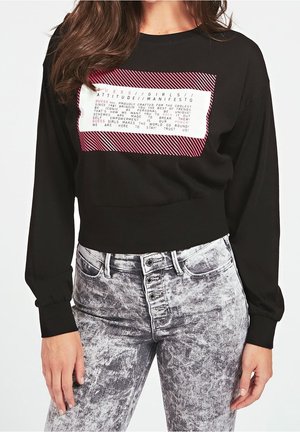 Femme portant un sweat-shirt court noir à manches longues avec un graphisme de texte blanc et rouge, associé à un jean gris taille haute délavé à l'acide.
