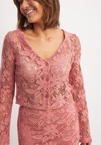 Pink lace cardigan with a V-neck, long sleeves, and decorative buttons. Features an open weave pattern with floral motifs and a cropped fit.