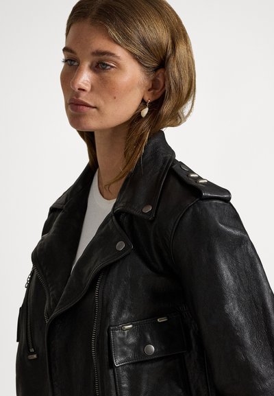 Crna kožna bajkerska jakna s epauletima, srebrnim ukrasima i džepovima na patentni zatvarač. Ima teksturiranu završnu obradu i široki dizajn ovratnika.