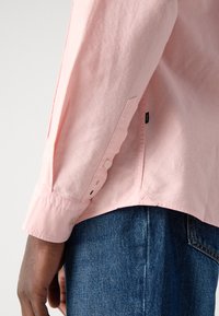Camicia button-down a maniche lunghe di un rosa chiaro, con polsini abbottonati e orlo curvo, abbinata a jeans in denim blu.