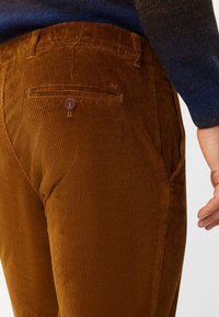 Braune Cordhosen mit vertikal gerippter Textur, einer Gesäßtasche mit braunem Knopfverschluss und seitlichen Taschen.