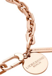 Bracelet en chaîne en or rose avec des maillons ovales, un fermoir et un médaillon circulaire gravé "LIEBESKIND BERLIN". Texture lisse et métallique.