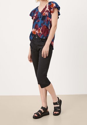 Femme portant un chemisier coloré à manches courtes et motifs abstraits, un pantalon noir court et des sandales noires ouvertes à l'avant avec des brides à la cheville.