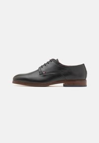 Chaussure de ville en cuir noir avec une finition lisse, bout arrondi, design à lacets et détails ton sur ton marron sur la semelle. Présente un logo à l'arrière.
