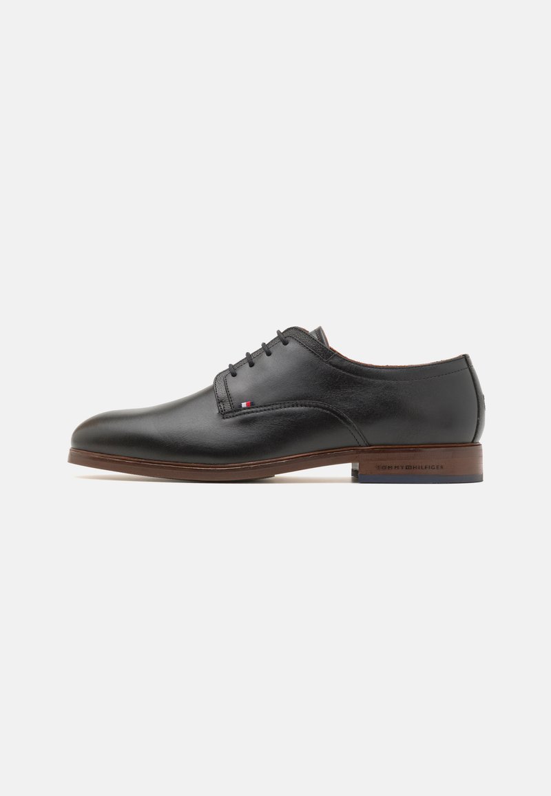 Chaussure de ville en cuir noir avec une finition lisse, bout arrondi, design à lacets et détails ton sur ton marron sur la semelle. Présente un logo à l'arrière.