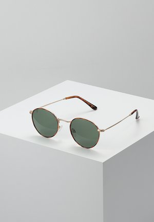 Gafas de sol redondas con lentes verdes, montura metálica plateada delgada y brazos con patrón de carey. Diseño minimalista sobre un fondo blanco.