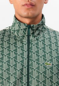 Zelená zip-up bunda s textúrovaným geometrickým vzorom v krémovej farbe. Má stojatý golier a malé vyšívané logo na hrudi.