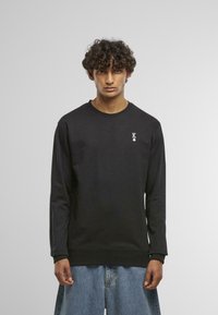 Melna garenīga krekla zupa ar apaļu kakla izgriezumu, ribotām aprocēm un mazu baltu "K" logotipu kreisajā krūtī. Mīksta, gluda tekstūra.