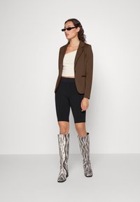 Hnědý přizpůsobený blazer, krémový zkrácený top, černé bikerské šortky a kozačky po kolena s vířivým černobílým vzorem. Doplňky: malé �černé sluneční brýle.