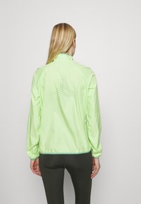 Chaqueta deportiva verde claro con cuello alto, puños elásticos y un detalle perforado en la parte posterior. Tejido suave y ligero.