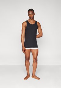 Black tank top with a round neckline, paired with white boxer shorts. The fabric has a smooth texture and fits closely to the body.