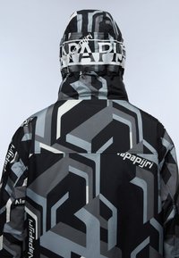 Giacca nera e grigia con motivo, dotata di cappuccio, che presenta forme geometriche astratte e un logo sul cappuccio, realizzata in materiale liscio e resistente all'acqua.
