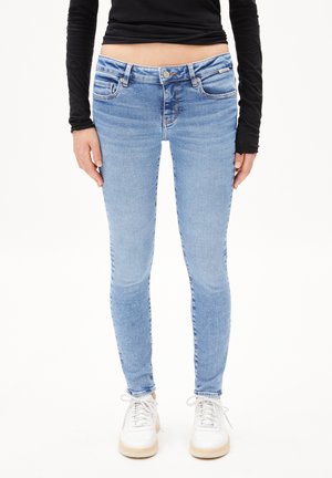 Jeans en coton bleu clair avec une coupe slim, cinq poches, un léger délavage, et une fermeture par bouton et zip. Portés avec des baskets blanches.