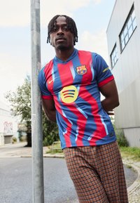 Raudonos ir mėlynos juostelės futbolo marškiniai su geltonu Spotify logotipu, dėvimi su rudomis languotomis kelnėmis. Modelis remiasi į stulpą miesto aplinkoje.