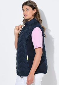Chaleco acolchado azul marino con un cuello alto, bolsillo lateral con cremallera y una camiseta corta de manga corta rosa de contraste por debajo. Pantalones blancos visibles.