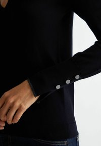 Zwarte gebreide trui met een V-hals, textuur aan de manchetten en drie grijze knopen op de mouw. Zacht materiaal met een getailleerde pasvorm.