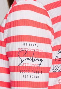 Striped long-sleeve top in white and coral, featuring ribbed texture. Design includes "Sunset Club" text and brand details in black.