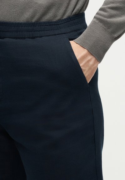 Χέρι στην πλαϊνή τσέπη ενός σκούρου ναυτικού παντελονιού με ελαστική μέση, φορεμένο με ανοιχτό γκρι υφασμένο πουλόβερ με μακριά μανίκια.