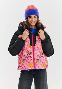 Ružová a čierna prešívaná bunda s vzorovaným predným dielom, ktorý obsahuje dizajn slnka, mesiaca a oka; obsahuje kapucňu a jasný zips. Modrá pletená čiapka.