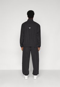 Survêtement noir comprenant une veste zippée avec col montant et poignets élastiques, associée à un pantalon ample assorti. Logo au dos de la veste.