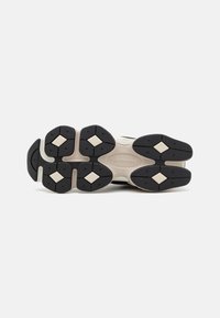 Semelle d'une chaussure New Balance montrant un motif en caoutchouc noir et beige avec des motifs de traction en forme de losange et le nom de la marque embossé au centre.