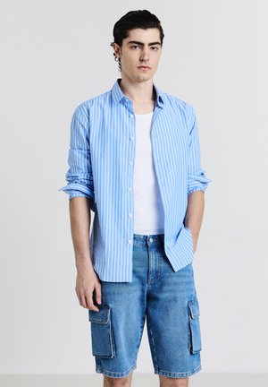 Giovane uomo che indossa una camicia a righe blu chiaro sopra una maglietta bianca e pantaloni cargo di jeans, in piedi con una mano in tasca.