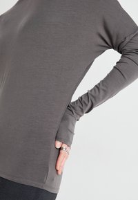 Long-sleeve gray top made of soft fabric, featuring a relaxed fit and side slits. Model’s hand is positioned on the hip, showcasing the design.