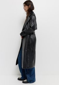 Femme portant un long trench-coat en cuir noir, un jean bleu à jambes larges et des chaussures noir et blanc, se tenant de profil contre un mur blanc.